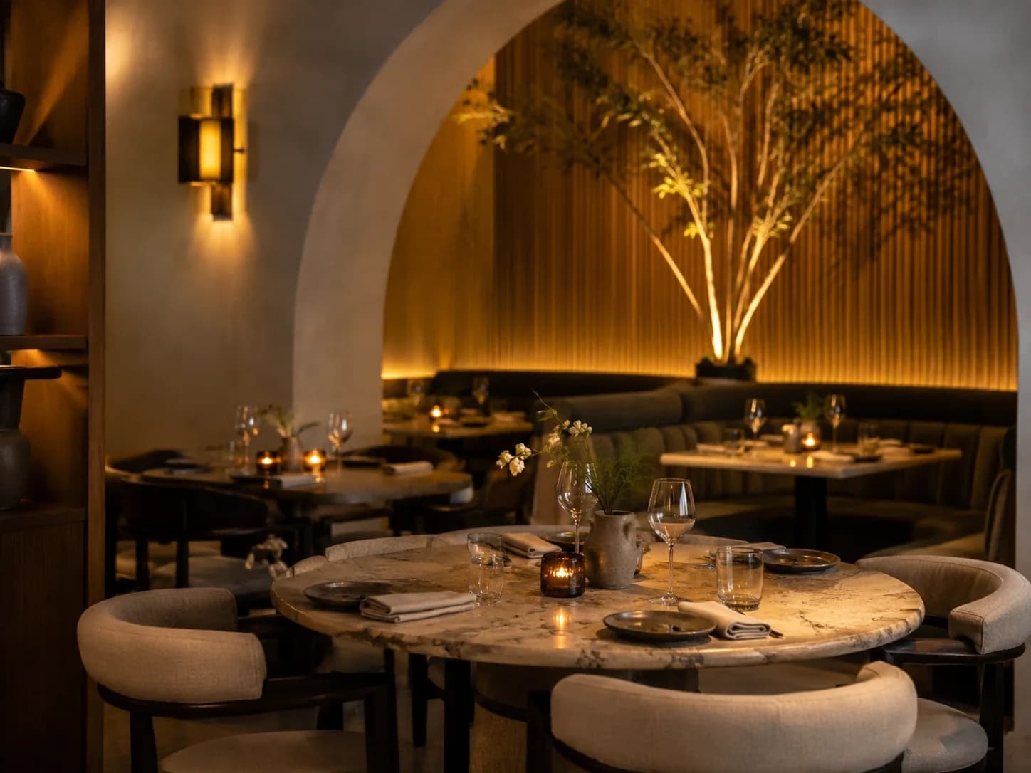 Candlelit restaurant room with a plaster arch, fluted timber wall, marble tables and an uplit tree.