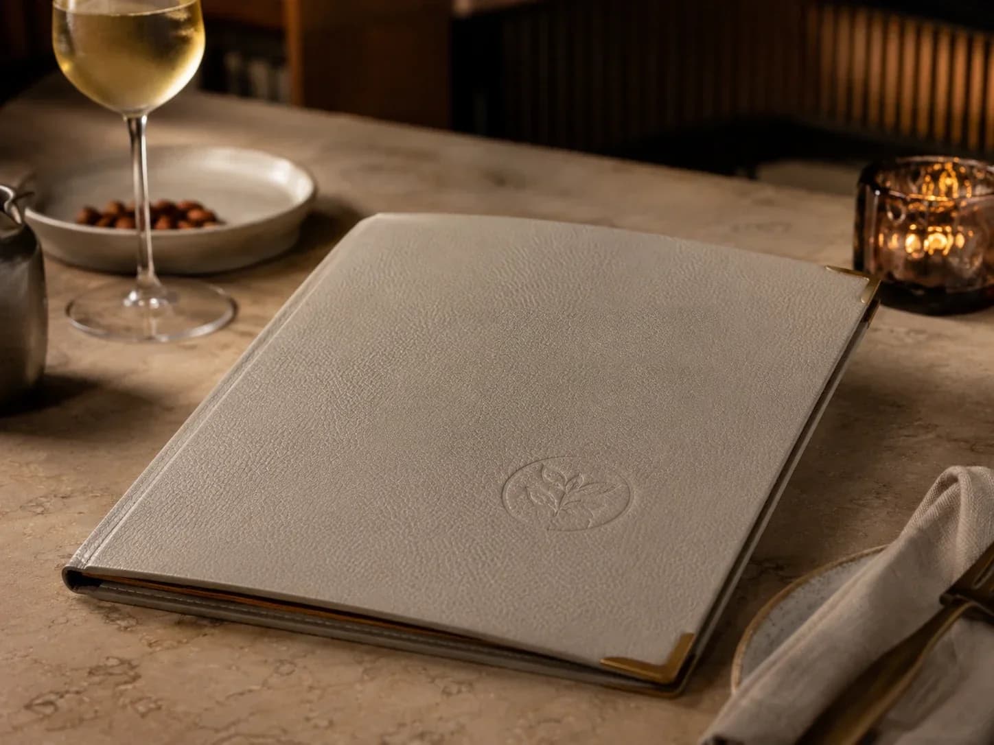 Cream leather menu folder embossed with a botanical sprig on a stone table beside a wine glass and candle.