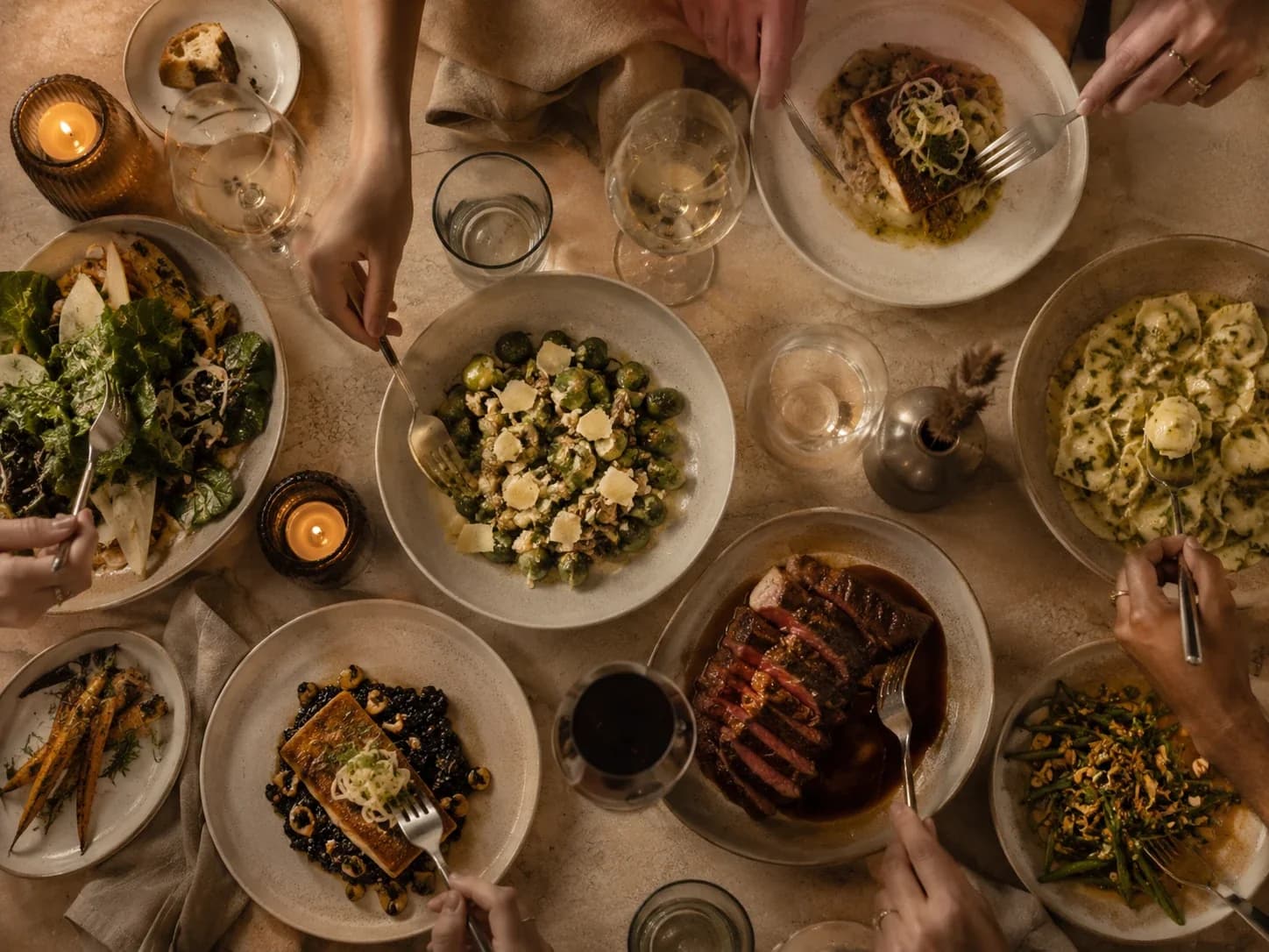 Overhead view of a shared stone table with several dishes, wine and hands reaching in to serve.