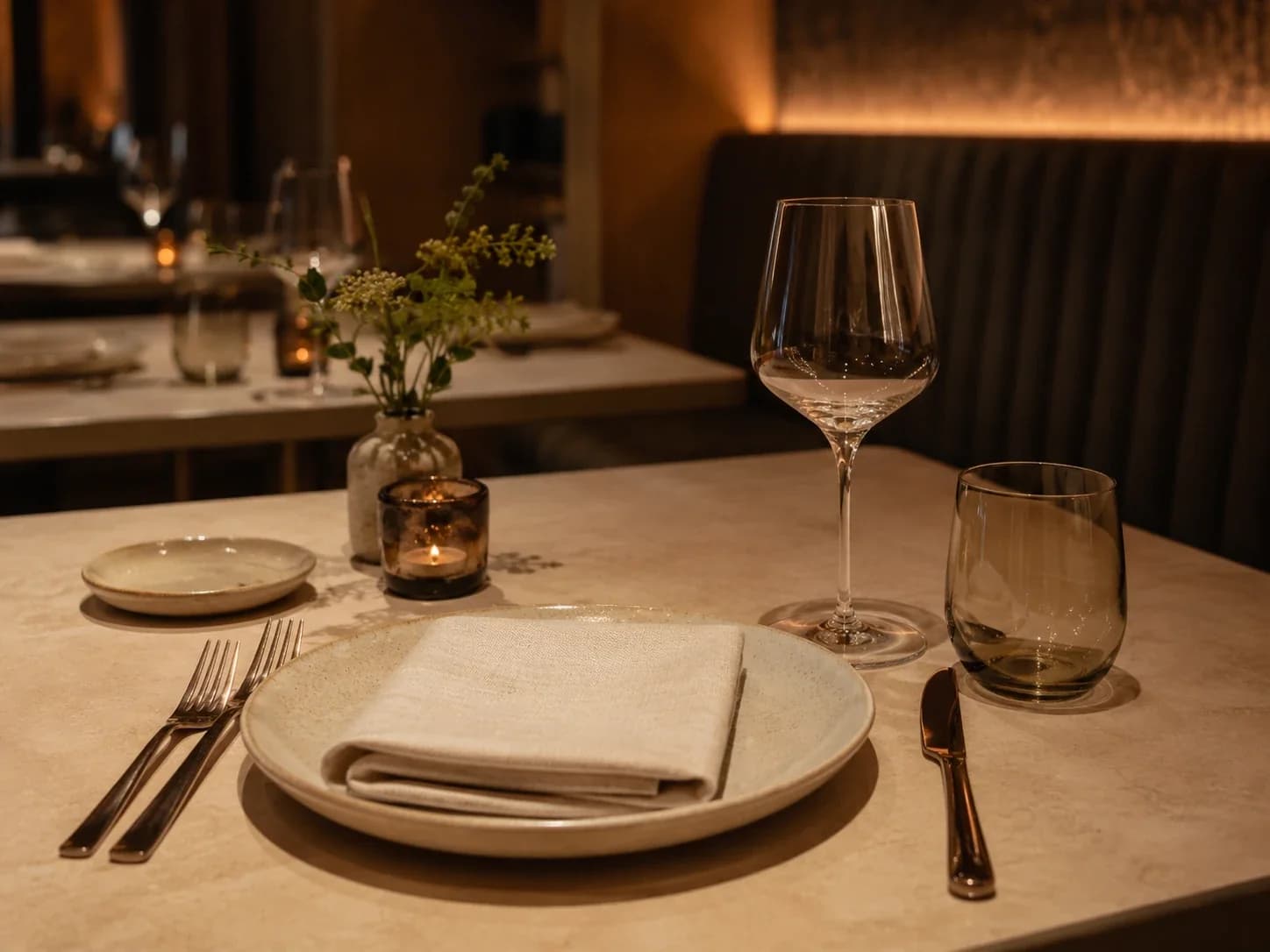 Candlelit table setting with a folded linen napkin on a speckled plate, wine glass, tumbler and a small vase of blossom.