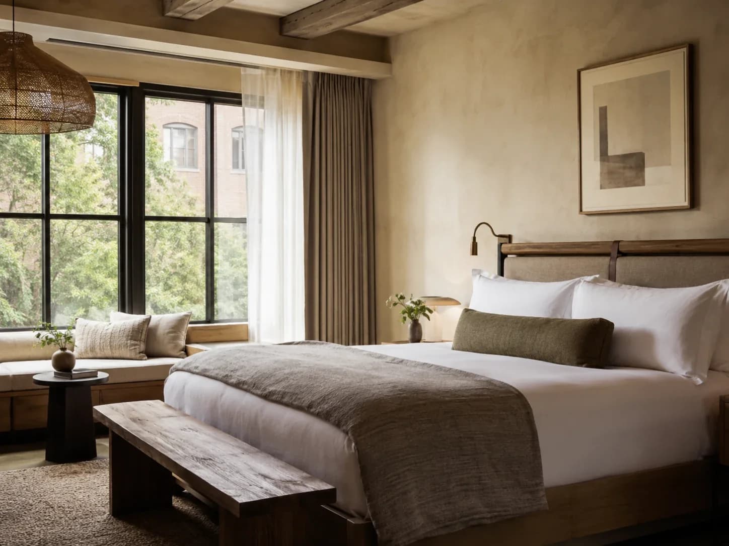 Boutique hotel bedroom with a linen-dressed bed, timber bench and a black-framed window onto trees.
