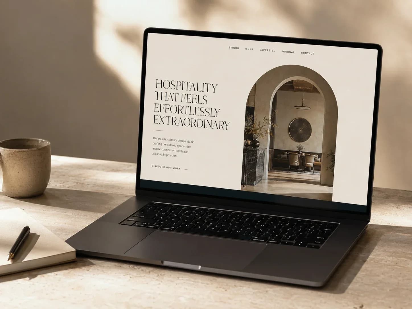 Laptop on a travertine desk showing an unbranded hospitality website design with an arched interior photograph.